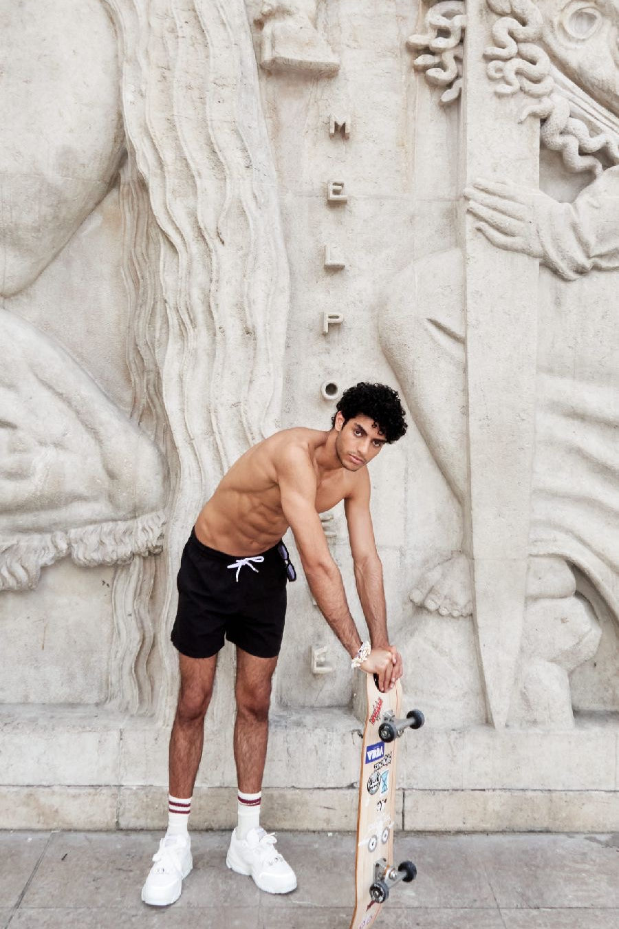 Photo d'un homme portant le short de bain noir. Il est torse nu et se tient de debout avec une planche de skate.