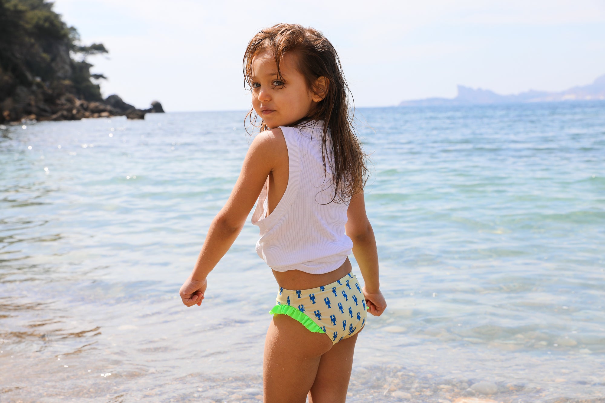 Photo d'une petite fille à la mer de dos portant la culotte de maillot de bain Lolo avec des homards bleus et des froufrous verts sur les côtés