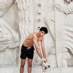 Photo d'un homme portant le short de bain noir. Il est torse nu et se tient de debout avec une planche de skate.
