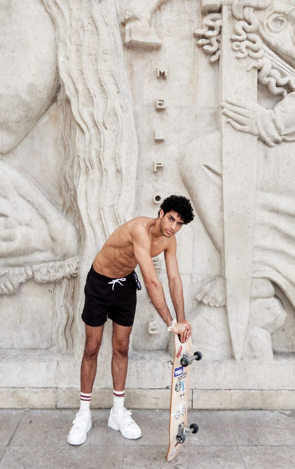 Photo d'un homme portant le short de bain noir. Il est torse nu et se tient de debout avec une planche de skate.