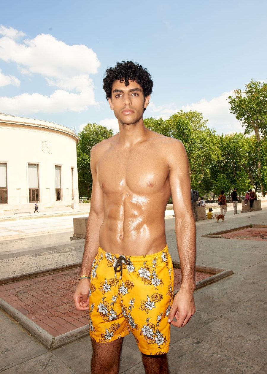 Photo d'un homme portant le short de bain jaune à fleur blanche. Il est torse nu et se tient de debout.